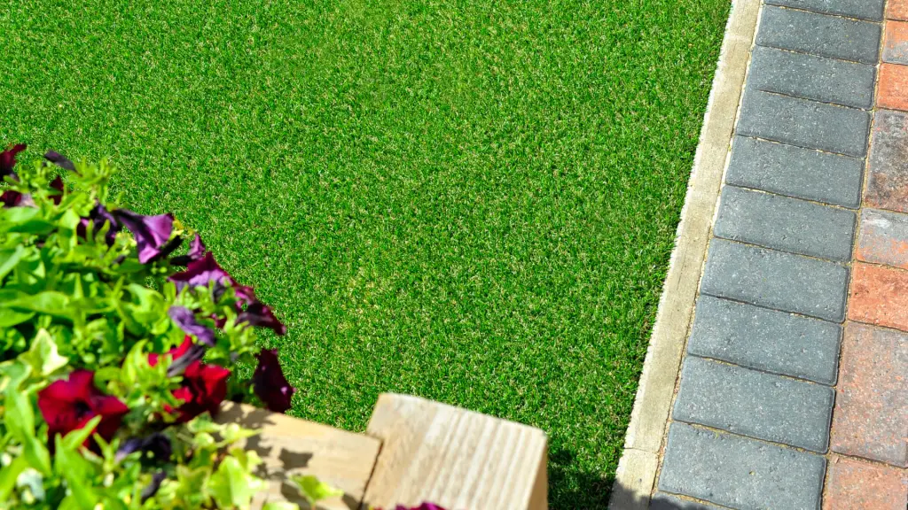 Borda de relva artificial ao lado de um canteiro com flores e um caminho de paralelepípedos